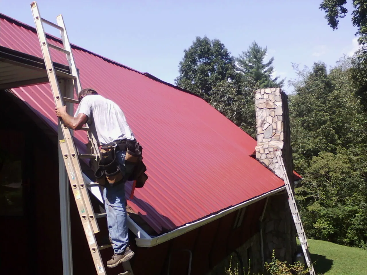 Roofer on a ladder beside a red metal roof and stone chimney — Soffit & Fascia Repair by Roofing Brothers in Connoquenessing
